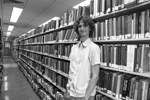 Stuart McMillen publicity photo 2015 black and white landscape. Standing between library shelves, looking at viewer with hand in pocket.