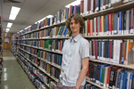 Stuart McMillen publicity photo 2015 colour landscape. Standing between library shelves, looking at viewer with hand in pocket.