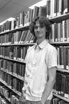 Stuart McMillen publicity photo 2015 black and white portrait. Standing between library shelves, looking at viewer with hand in pocket.
