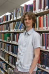 Stuart McMillen publicity photo 2015 colour portrait. Standing between library shelves, looking at viewer with hand in pocket.